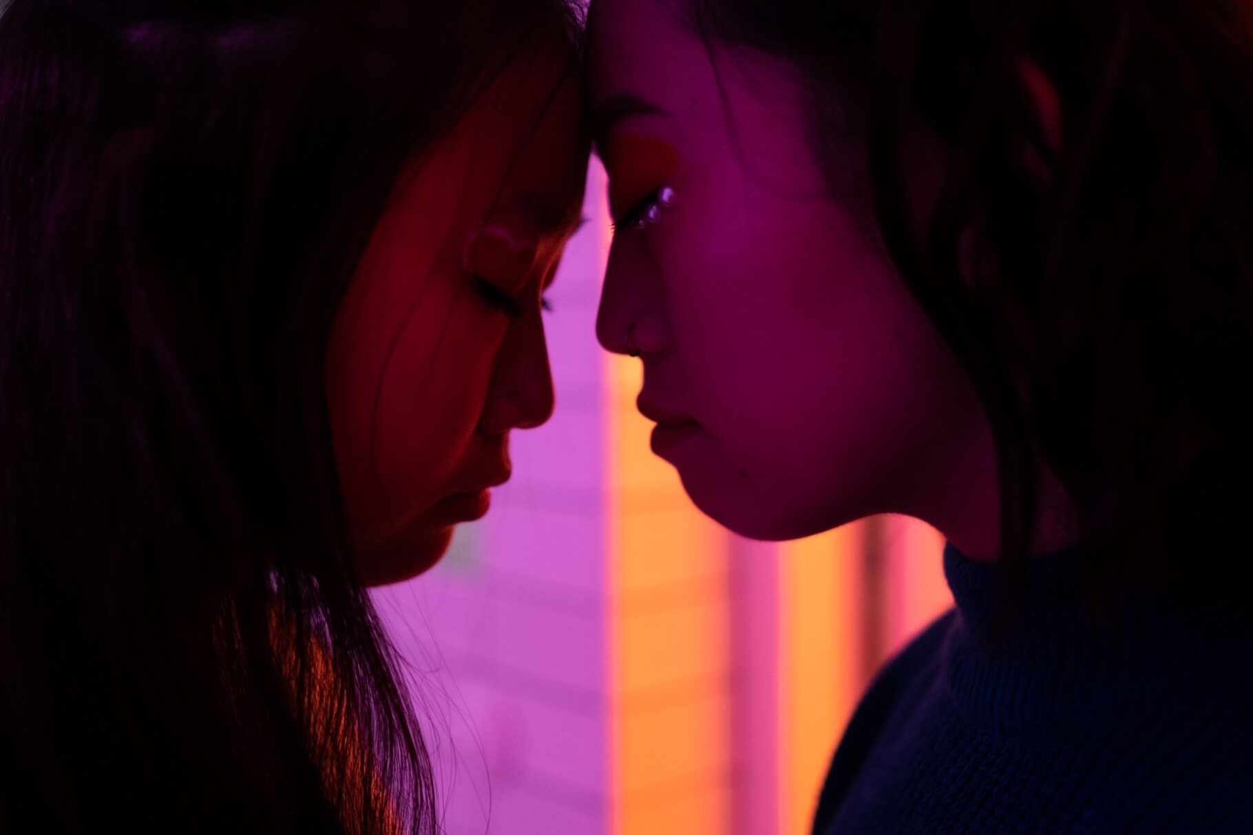 Intimate profile of two women touching foreheads bathed in vibrant neon light.